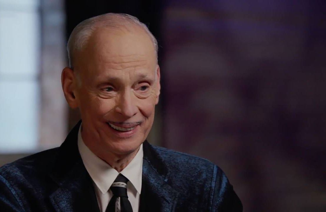 John Waters wearing a suit and tie, smiling. Next Avenue,