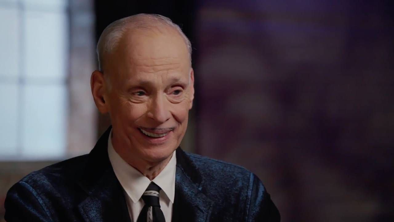 John Waters wearing a suit and tie, smiling. Next Avenue,