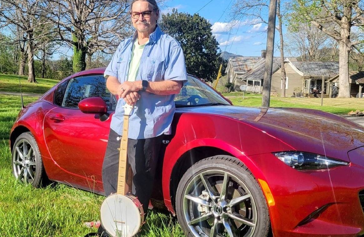 A man standing in front of a red car while holding a banjo. Next Avenue, volunteering in retirement