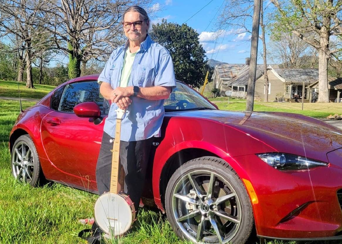A man standing in front of a red car while holding a banjo. Next Avenue, volunteering in retirement