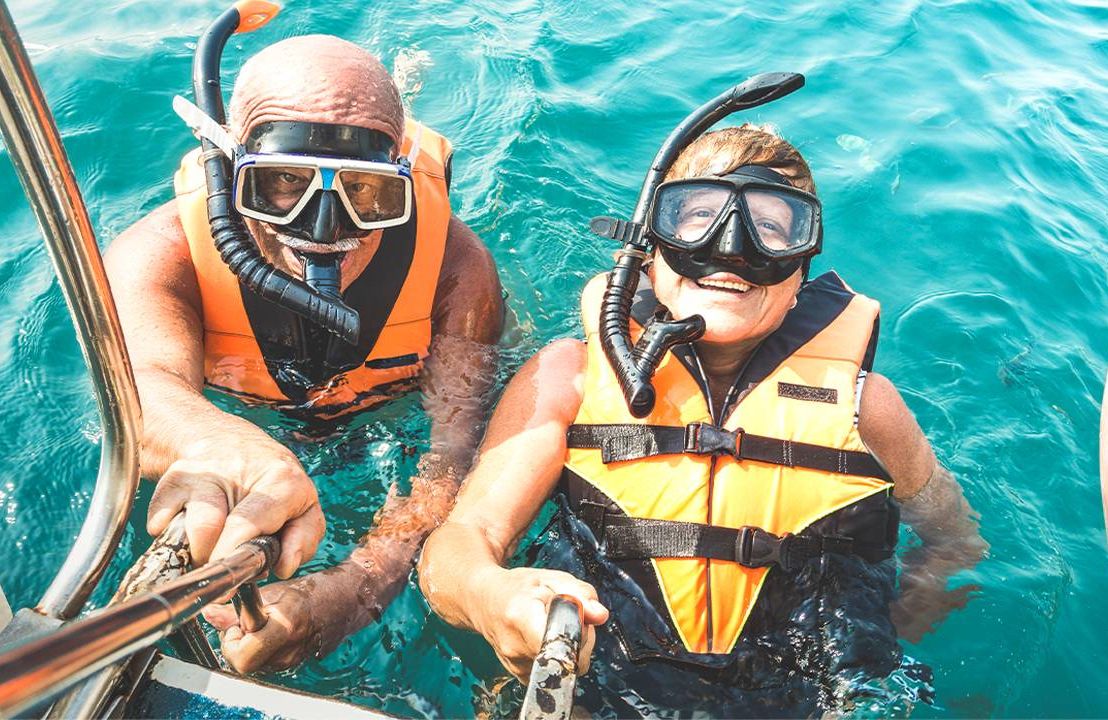 Two older adults wearing snorkeling gear in the ocean. Next Avenue, is travel insurance worth it