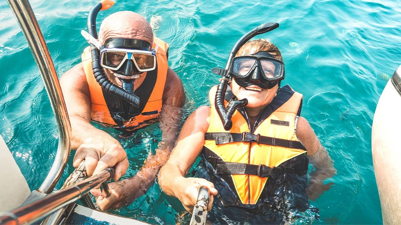 Two older adults wearing snorkeling gear in the ocean. Next Avenue, is travel insurance worth it