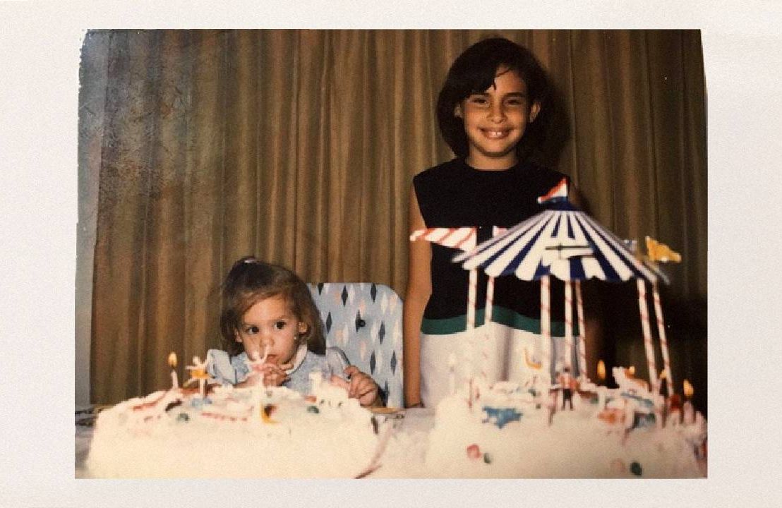 An old photo of two young girls standing in front of two birthday cakes. Next Avenue, sisters