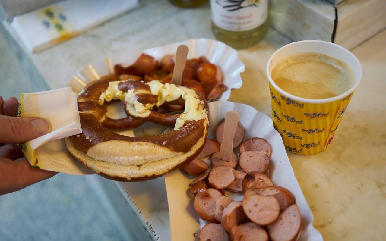 A person holding a pretzel with currywurst sausages in the background. Next Avenue, gluten free food tour