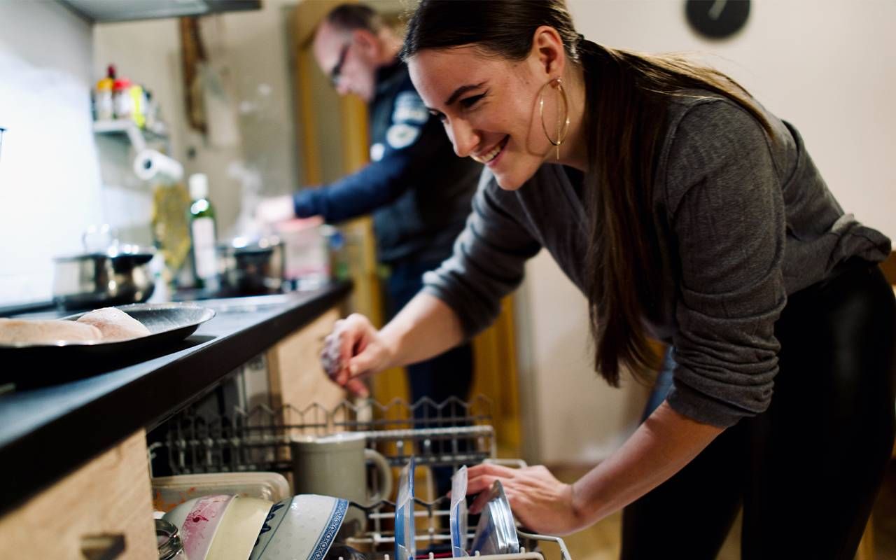 A young woman loading a dishwasher while her dad cooks a meal. Next Avenue, moving back home, living with parents