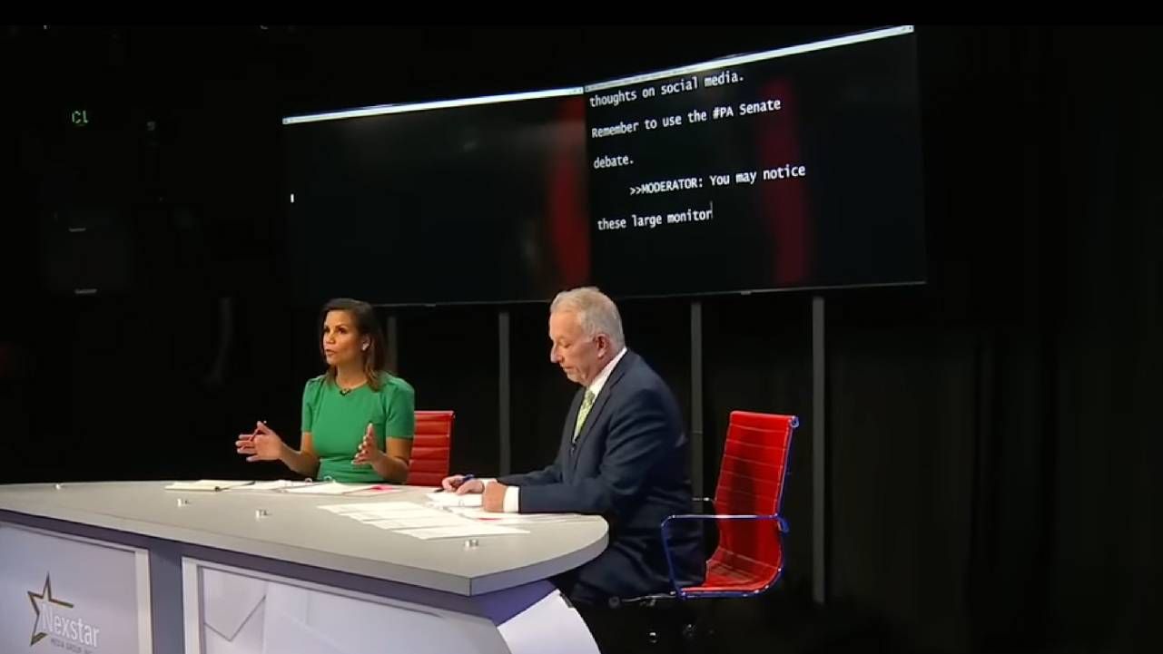 Two debate hosts with two large TV monitors displaying closed captions. Next Avenue, pennsylvania senate debate, stroke