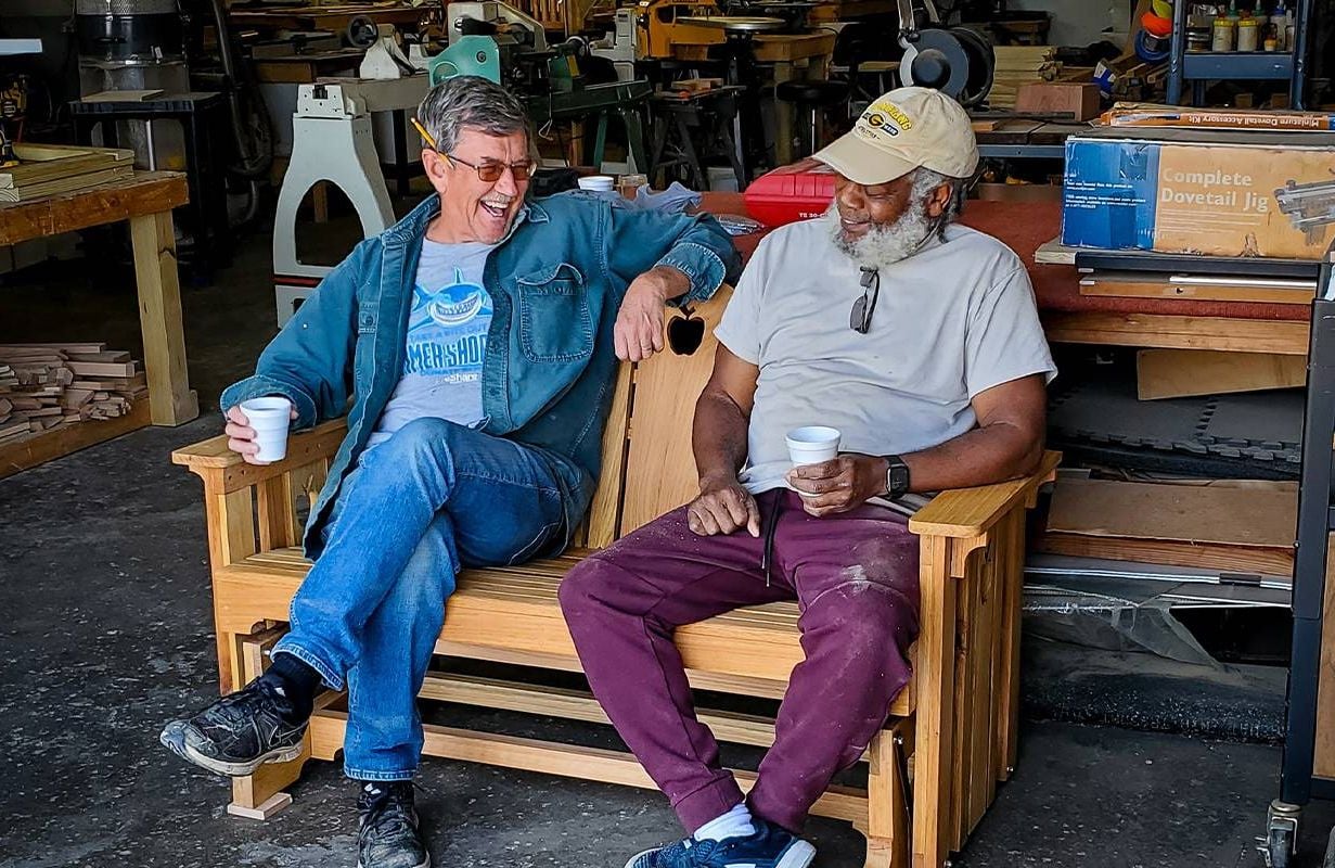 Two men sitting on a wooden bench in a shed. Next Avenue, men's shed