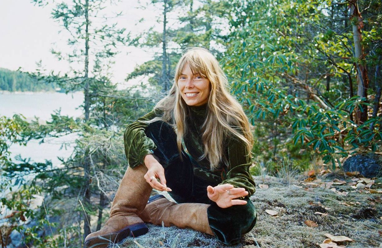 Joni Mitchell sitting on a rock in the woods overlooking a lake. Next Avenue