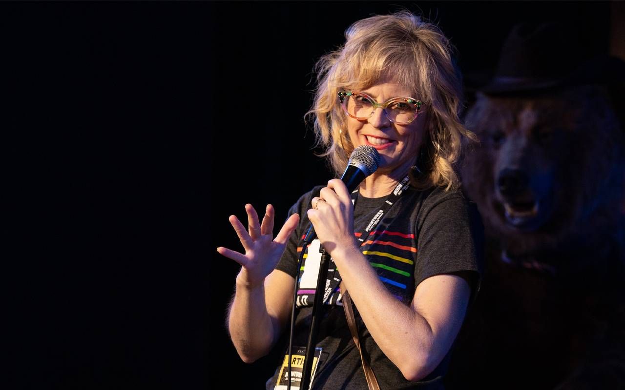 A woman on stage holding a mic during a comedy show. Next Avenue