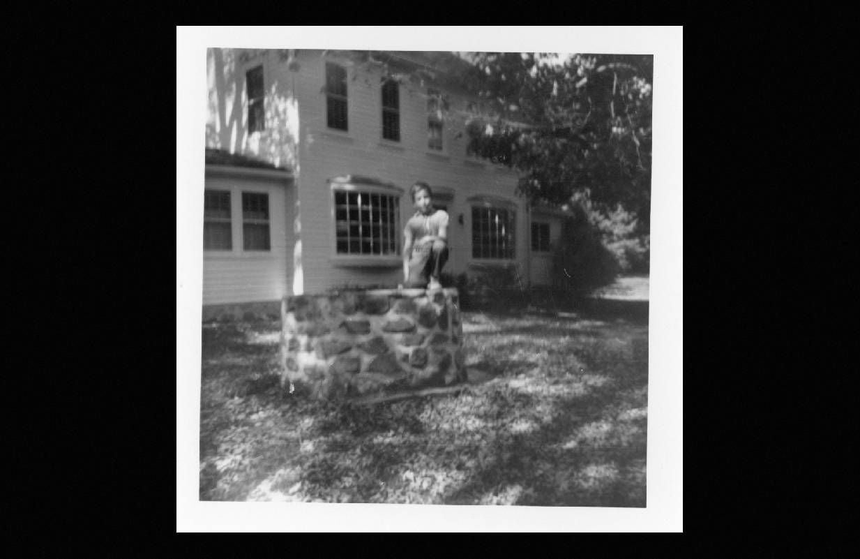 A black and white photo of a young boy sitting outside of a big white house. Next Avenue