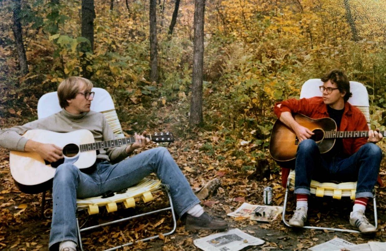 An old photo of two people sitting in the woods playing guitar together. Next Avenue