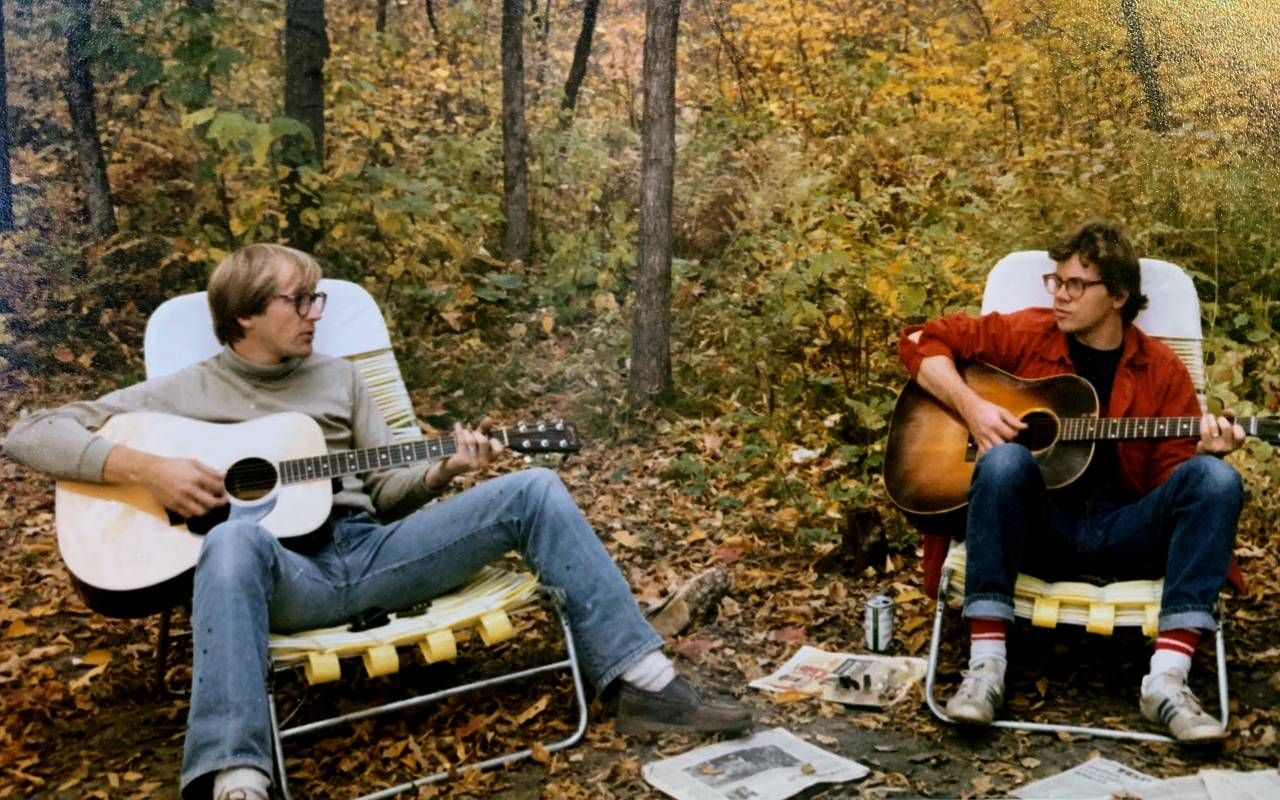 An old photo of two people sitting in the woods playing guitar together. Next Avenue