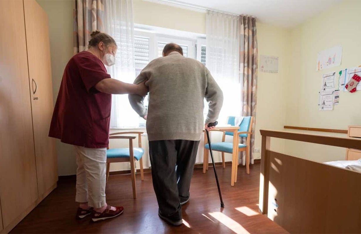 A staff member at a nursing home helping a patient into a chair. Next Avenue