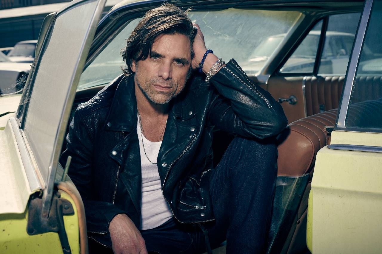 A man wearing a leather jacket sitting in a vintage car. Next Avenue, John Stamos