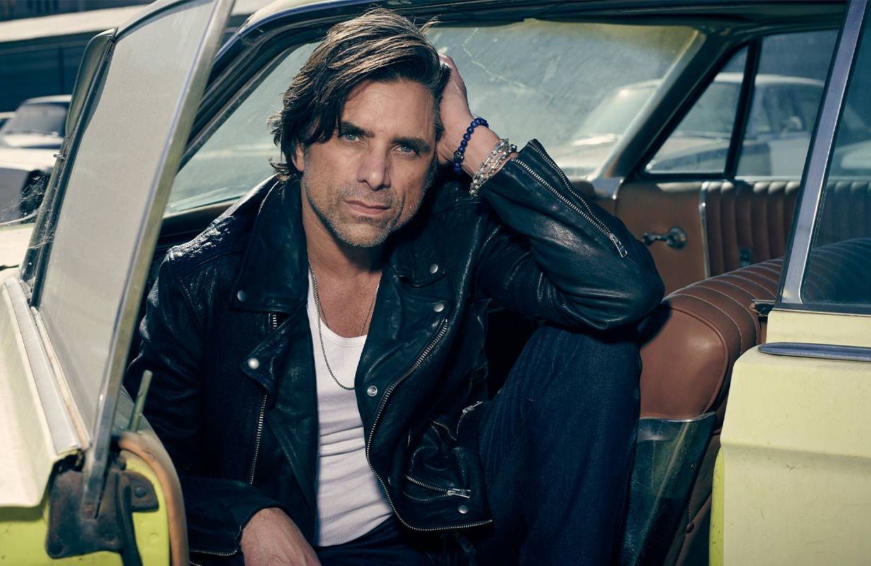 A man wearing a leather jacket sitting in a vintage car. Next Avenue, John Stamos