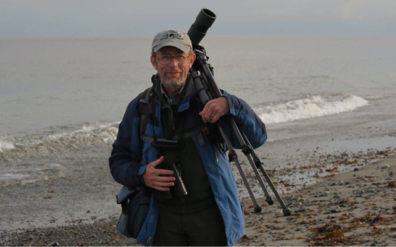 A man standing on a beach holding a tripod and camera. Next Avenue