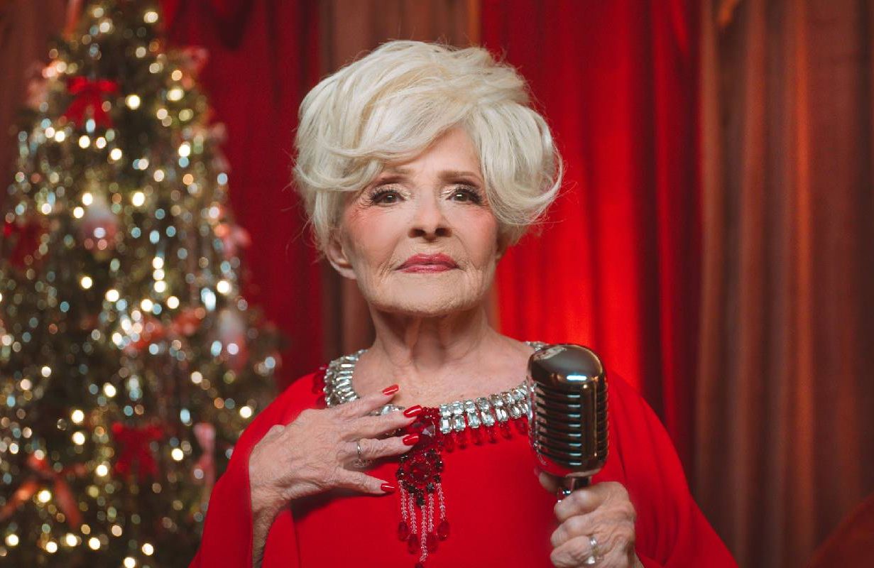 Brenda Lee wearing a red dress and holding a microphone in front of a Christmas tree. Next Avenue