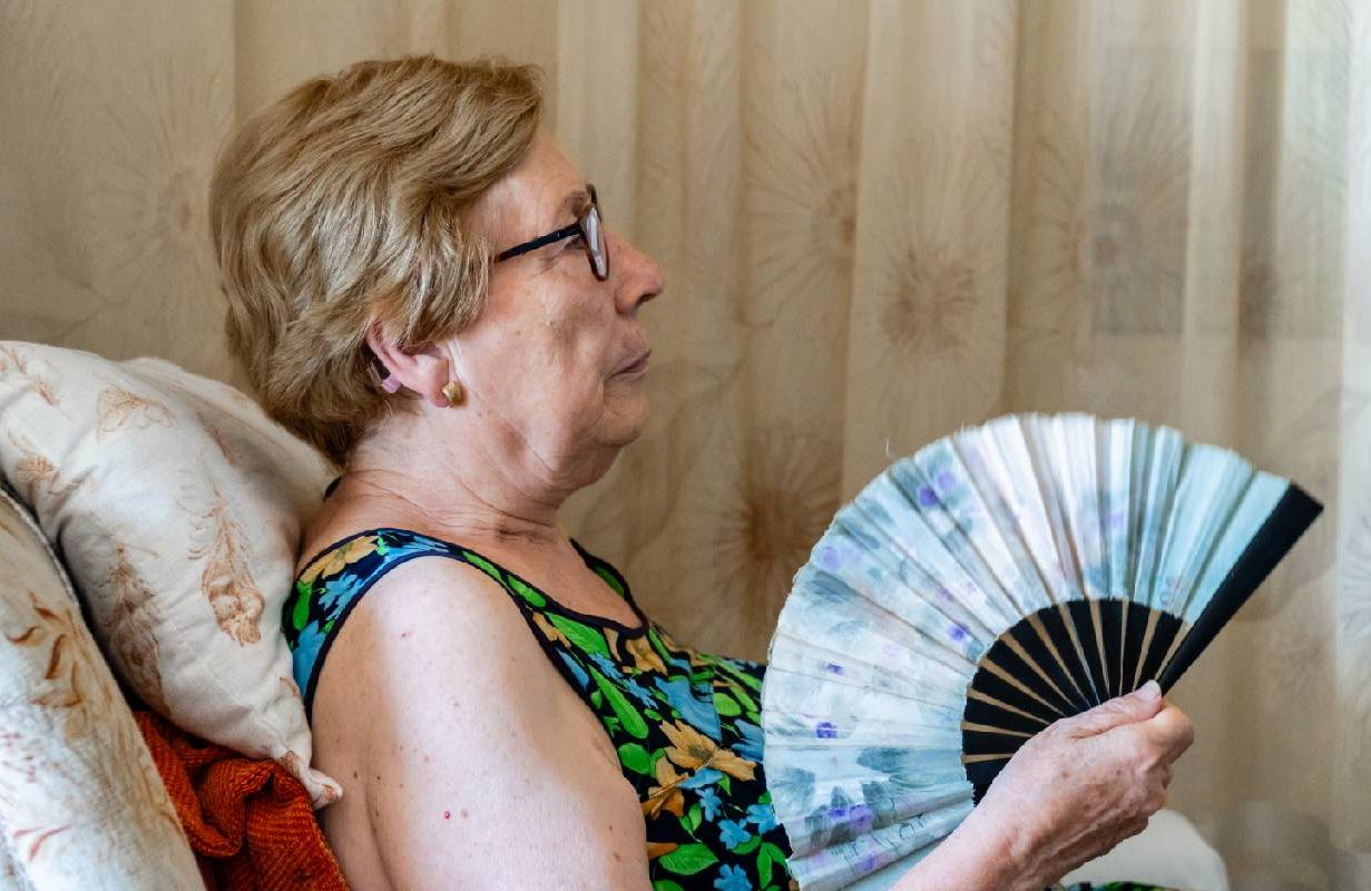 A woman who is sweating and holding a fan at home. Next Avenue
