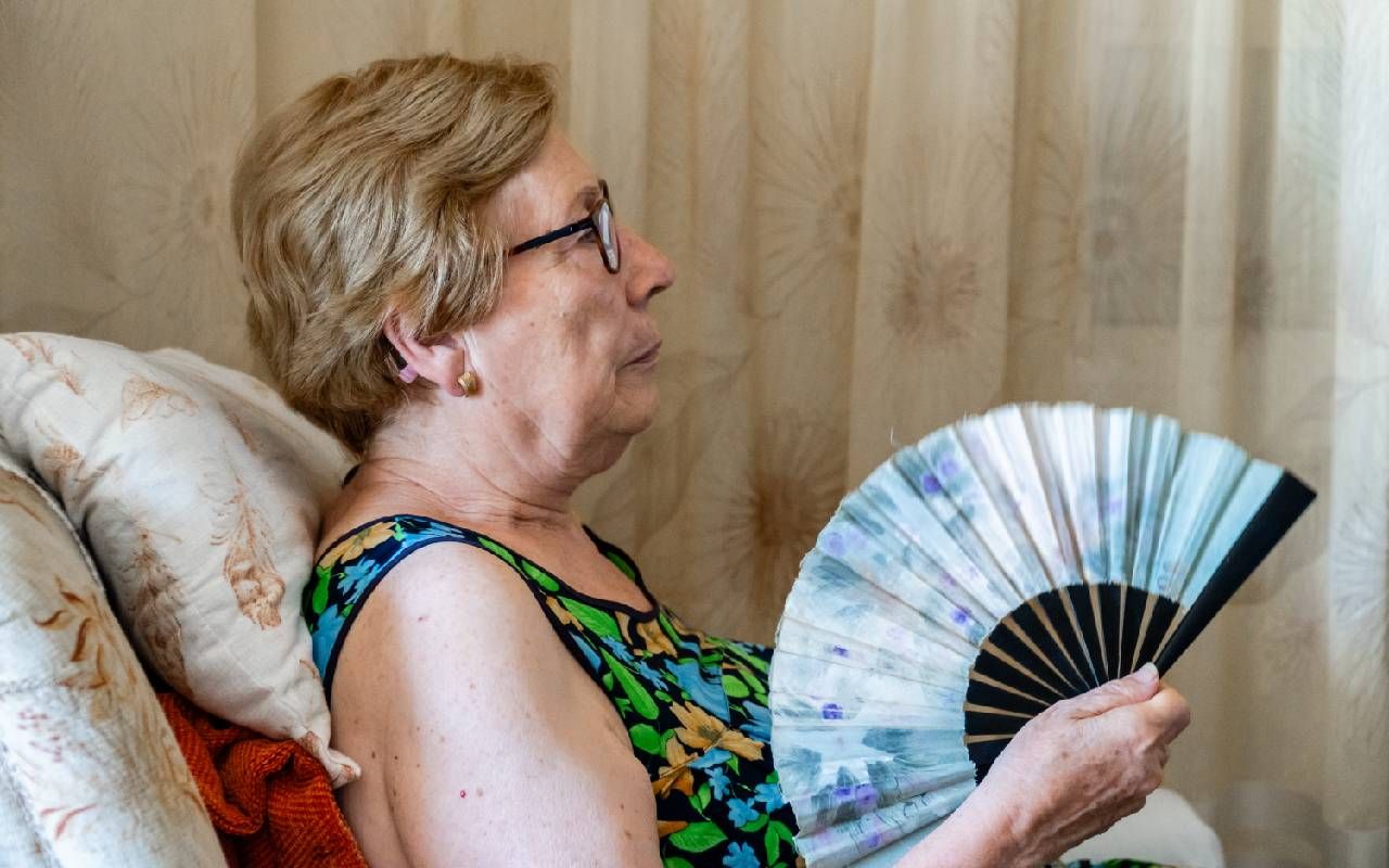 A woman who is sweating and holding a fan at home. Next Avenue