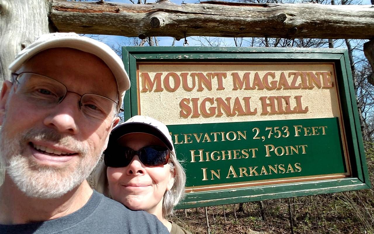 A couple smiling in front of a sign at the base of a mountain. Next Avenue