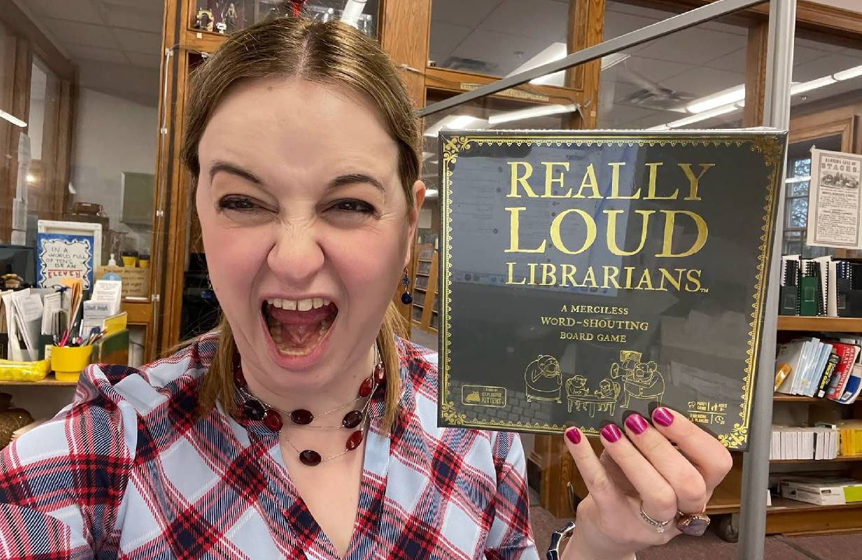 A person with a screaming look on their face holding up a board game titled, "Really Loud Librarians". Next Avenue