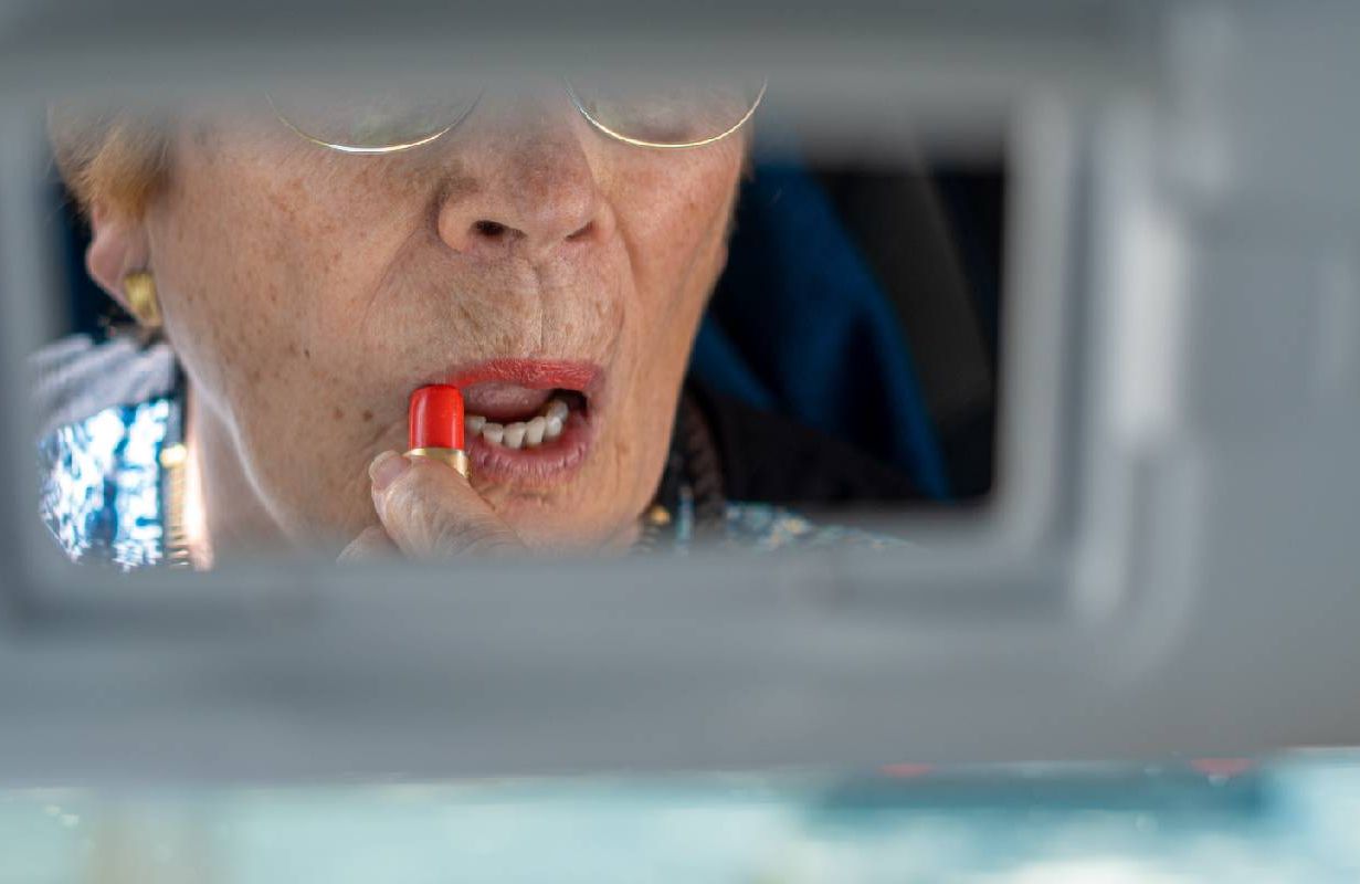 A older woman putting on lipstick in her car's rear view mirror. Next Avenue