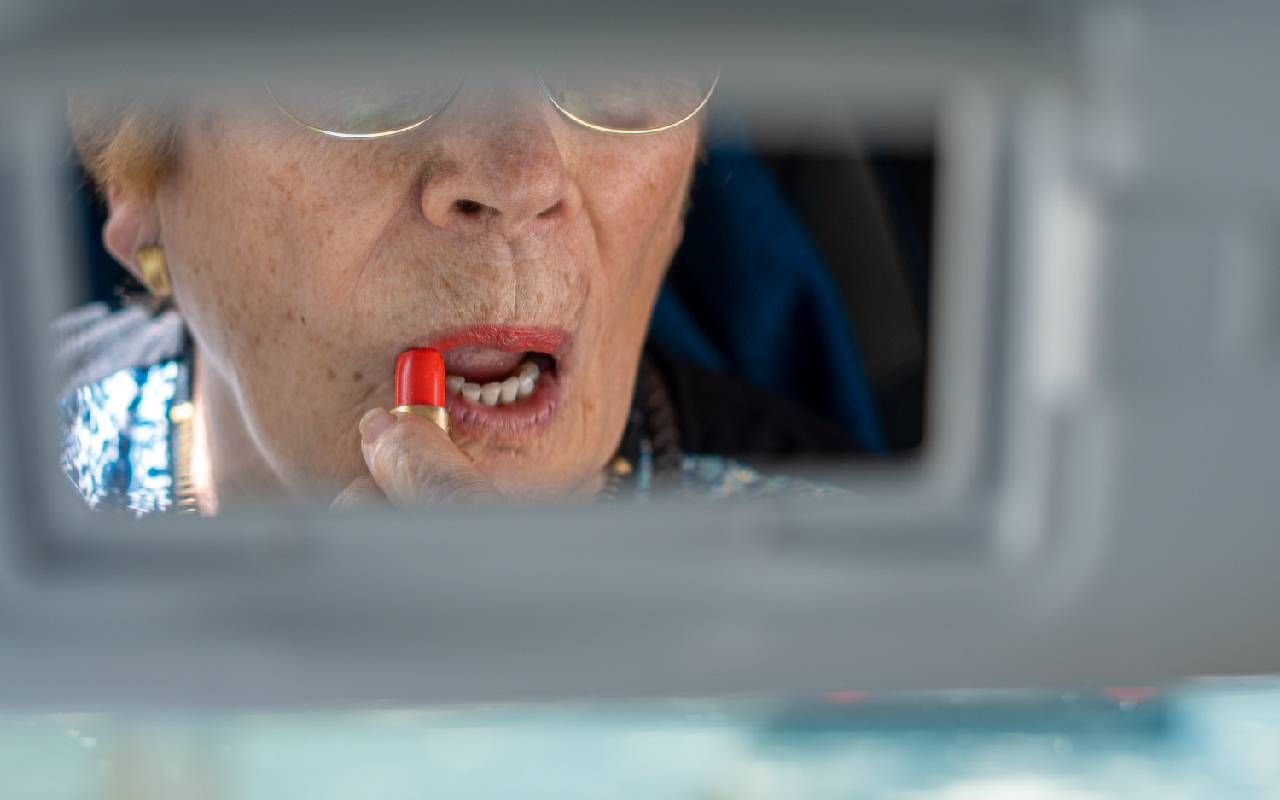 A older woman putting on lipstick in her car's rear view mirror. Next Avenue