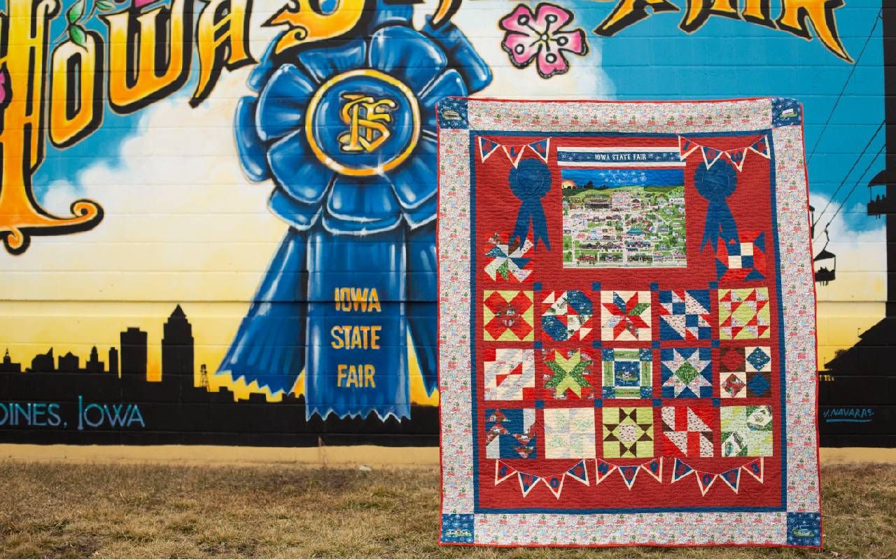 A quilt held up in front of an Iowa State Fair mural. Next Avenue, quilting