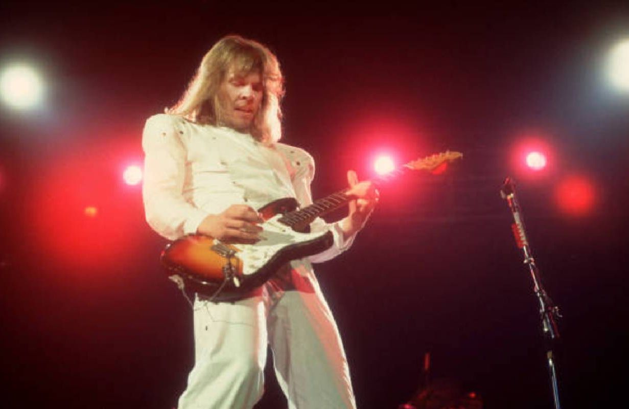 James Young wearing a white suit playing guitar on stage with Styx. Next Avenue