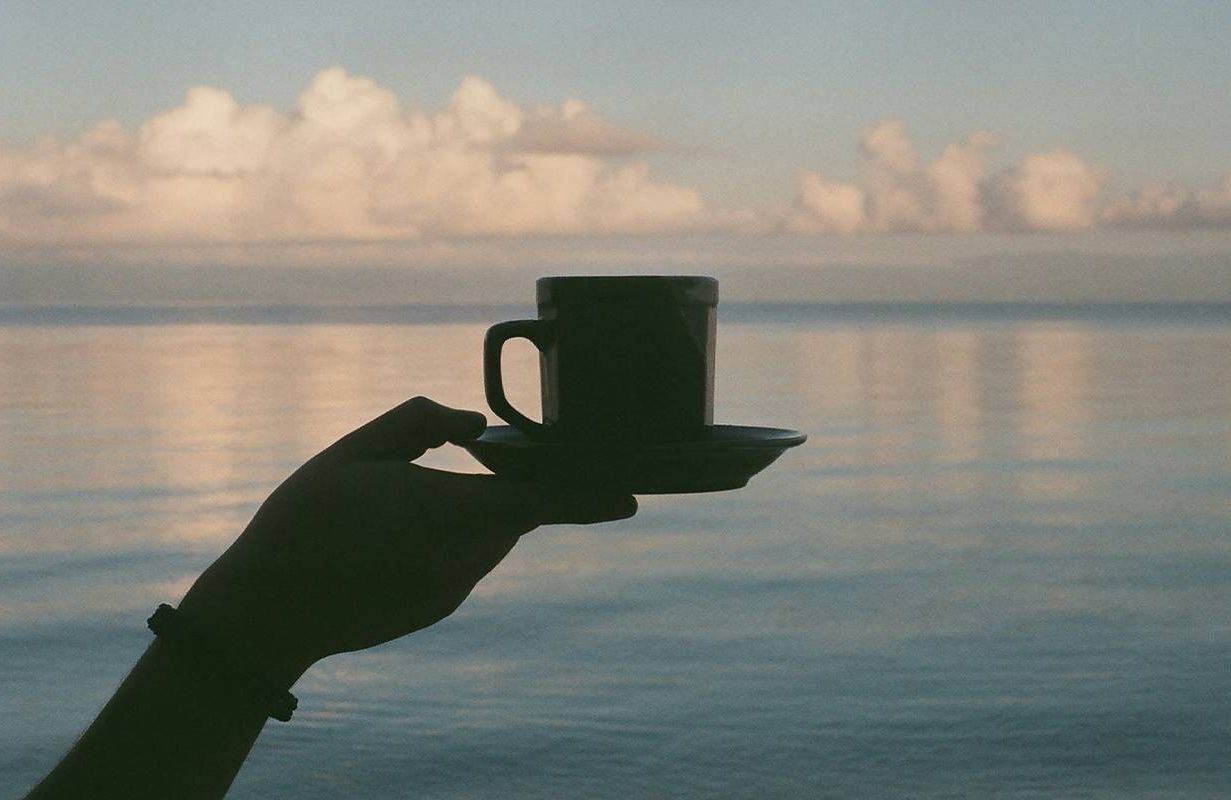A person holding a cup of coffee with the ocean in the background. Next Avenue, caffeine, pros and cons