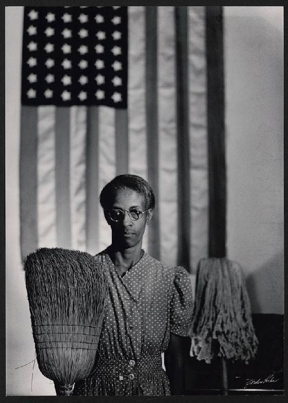 A black and white photograph of A womam mholding a broom and a mop in front of the American flag. Next Avenue, Gordon Parks, Photography