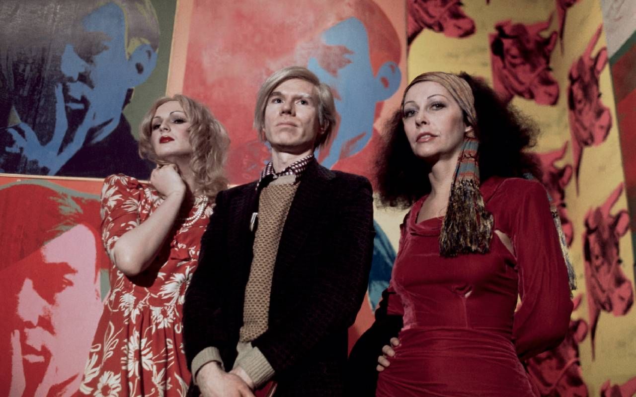 Andy Warhol posing with two people in front of colorful art. Next Avenue, Candy Darling