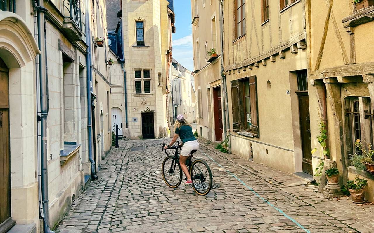 A woman riding a bike in down a cobblestone street in France. Next Avenue, older adults learning another language