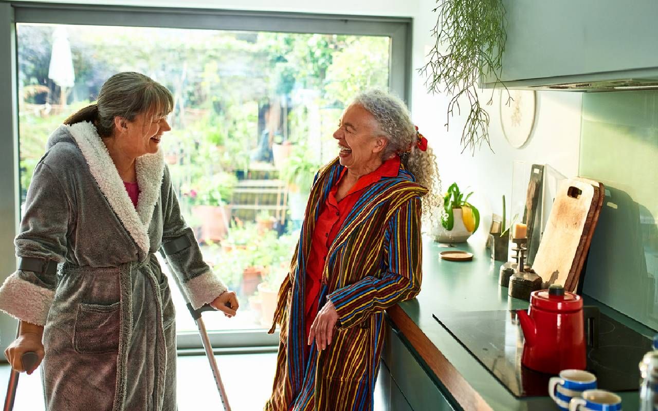 Two older roommates or boommates laughing together in the kitchen. Next Avenue