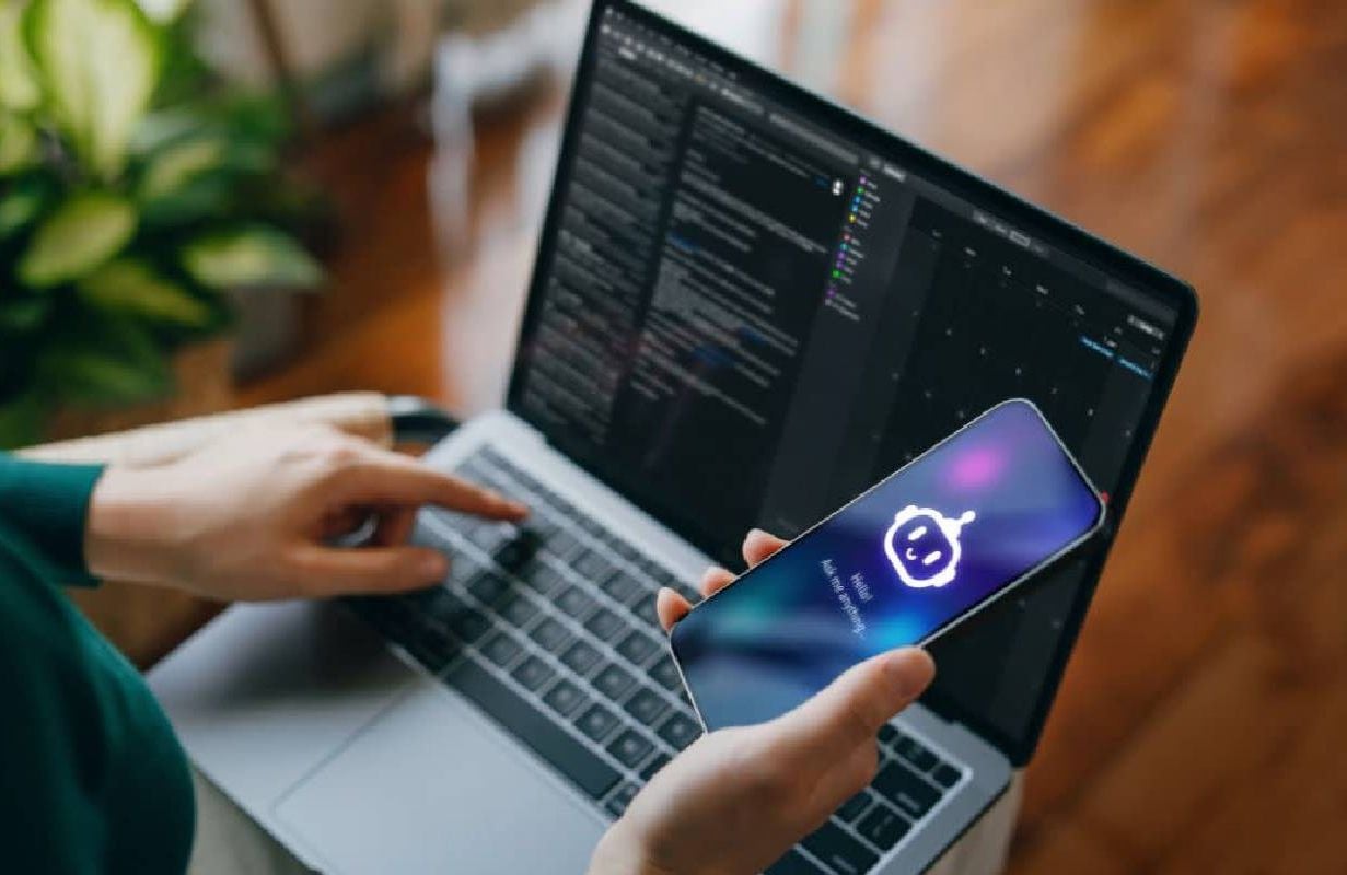 Woman using AI chatbot virtual assistance app on smartphone to assist her while working on laptop. Next Avenue