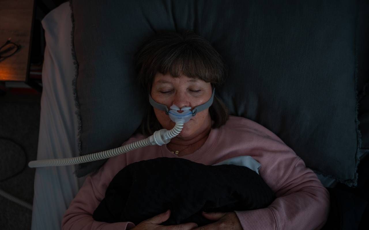 A person sleeping in bed wearing a CPAP machine mask. Next Avenue, chronic fatigue