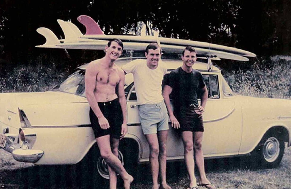An old photo of three people smiling in front of surfboards. Next Avenue, entrepreneurs