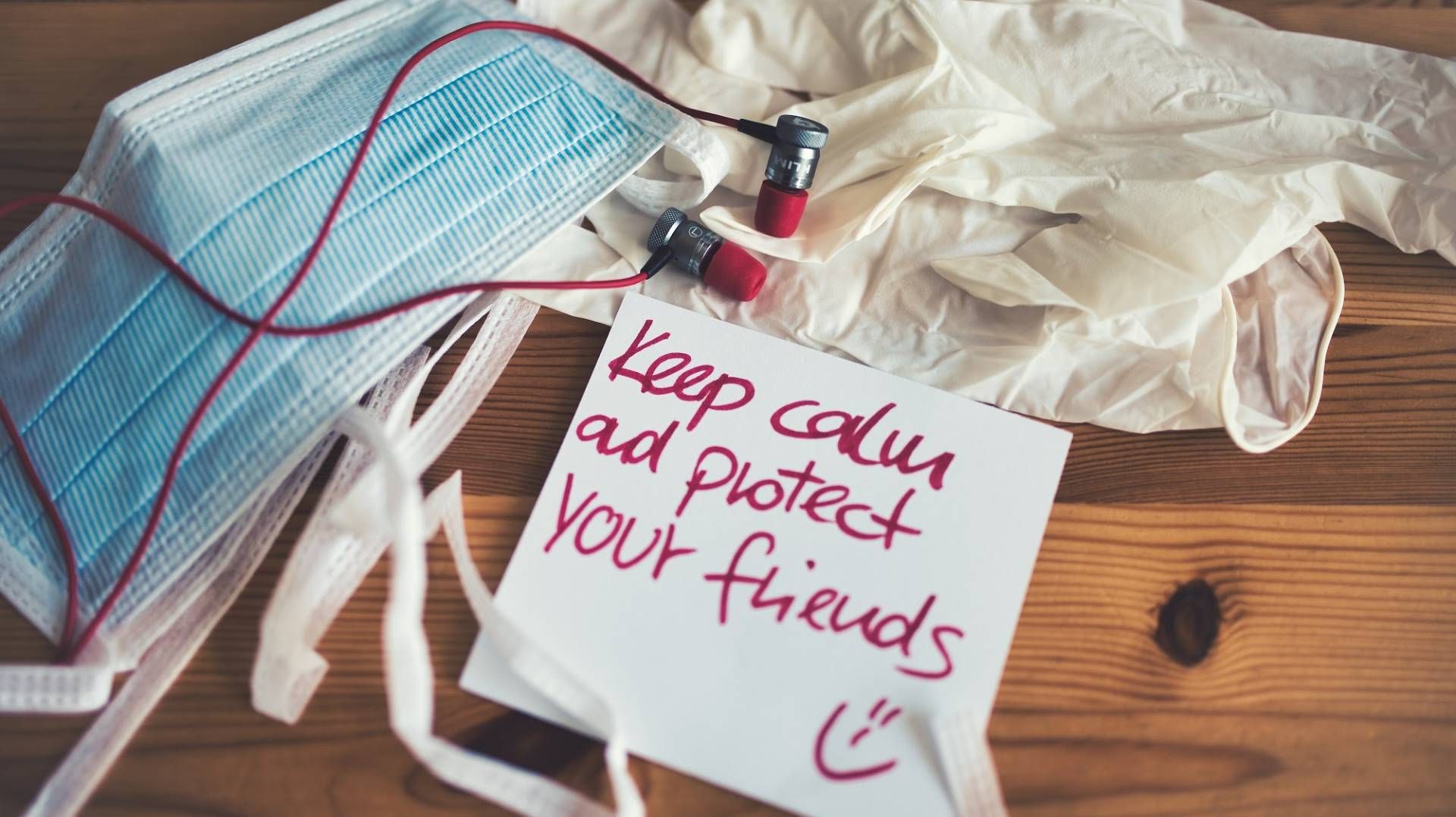 A blue surgical mask, a pair of medical gloves and some ear plugs are piled together on a wooden table.  Next to the items is a white, square note with "Keep Calm and protect your friends" and a smiley face is written in dark pink ink. 