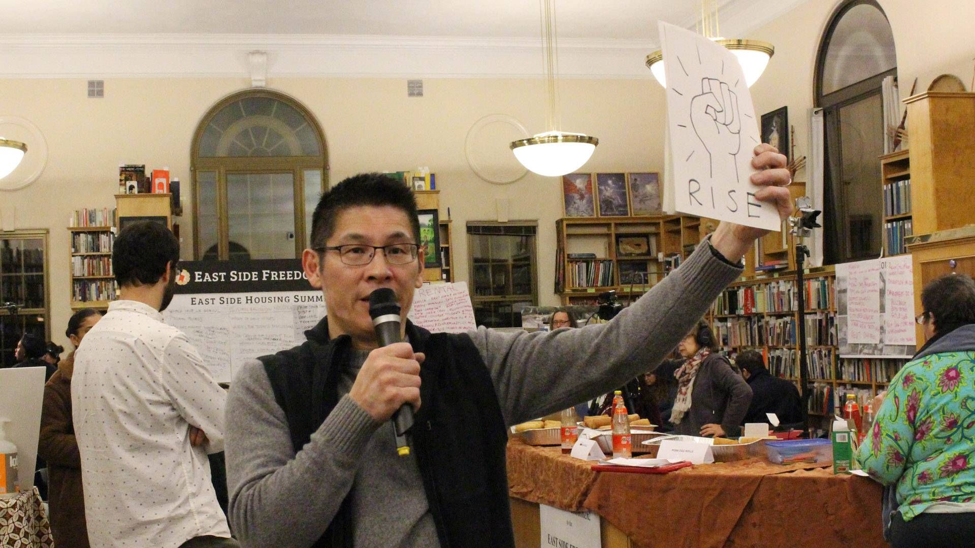 Asian man holds up a sign that reads rise inside of a library