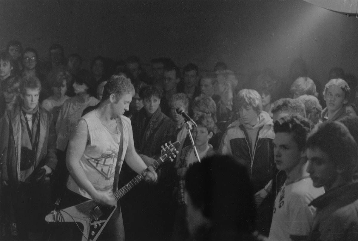 The band Hüsker Dü - one person with a guitar standing in front of a microphone - plays in front of a crowd of people dressed in 1980s clothing.