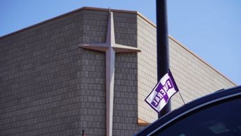 A funeral flag waves from atop an SUV in the funeral procession. A cross is fixed to the front of the church.