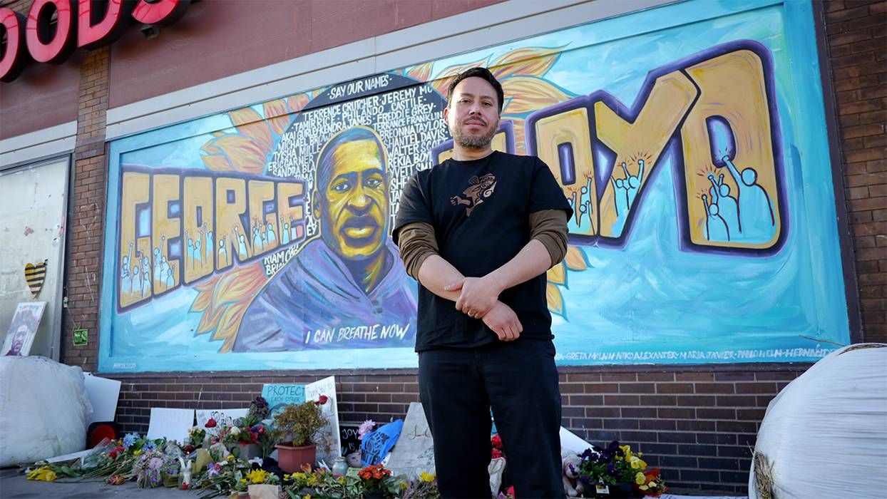 A man stands in front of a mural of George Floyd.