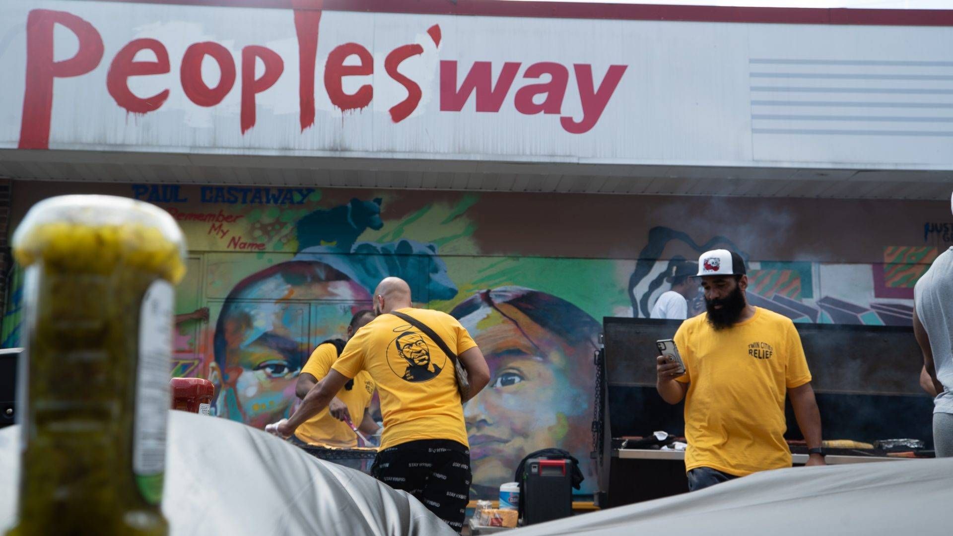 Members of the Twin Cities Relief initiative cook cheese burgers and hot dogs outside of a speedway gas station at George Floyd Square. The gas station has been painted over, reading "People's Way."