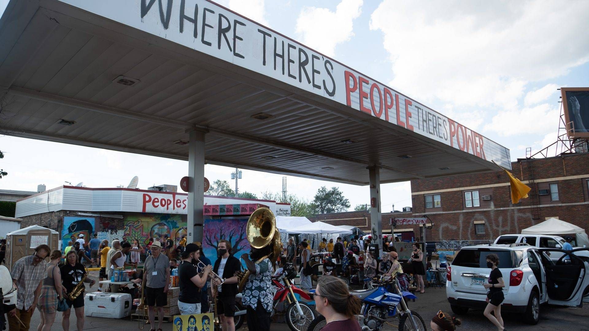Dozens of people gather under the Speedway gas station at George Floyd Square. The station is painted over, reading "Where there's people there's power"