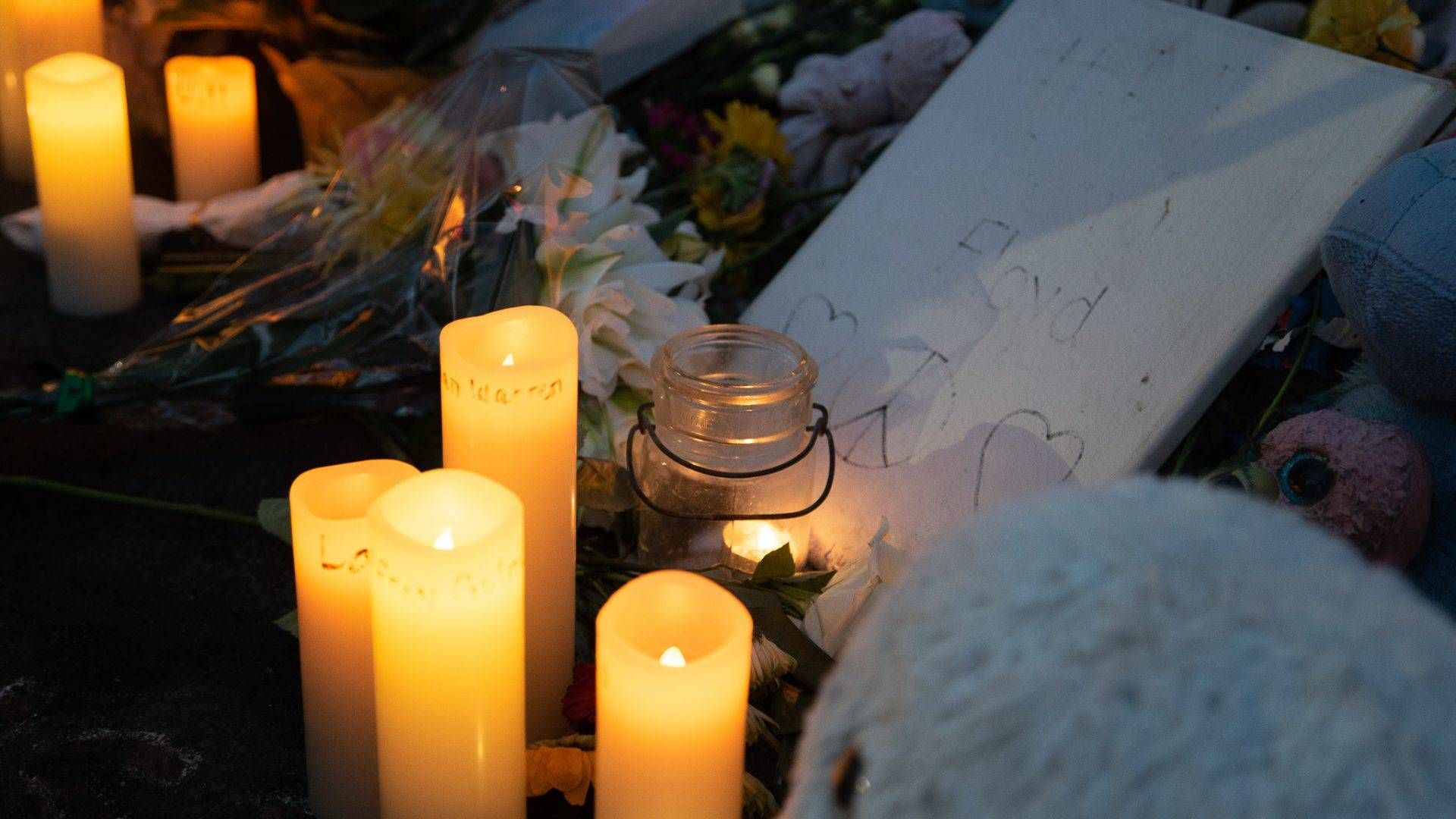 Candlelights placed near a teddy bear, flowers and a faded sign with George Floyd's name at a vigil commemorating Floyd's life