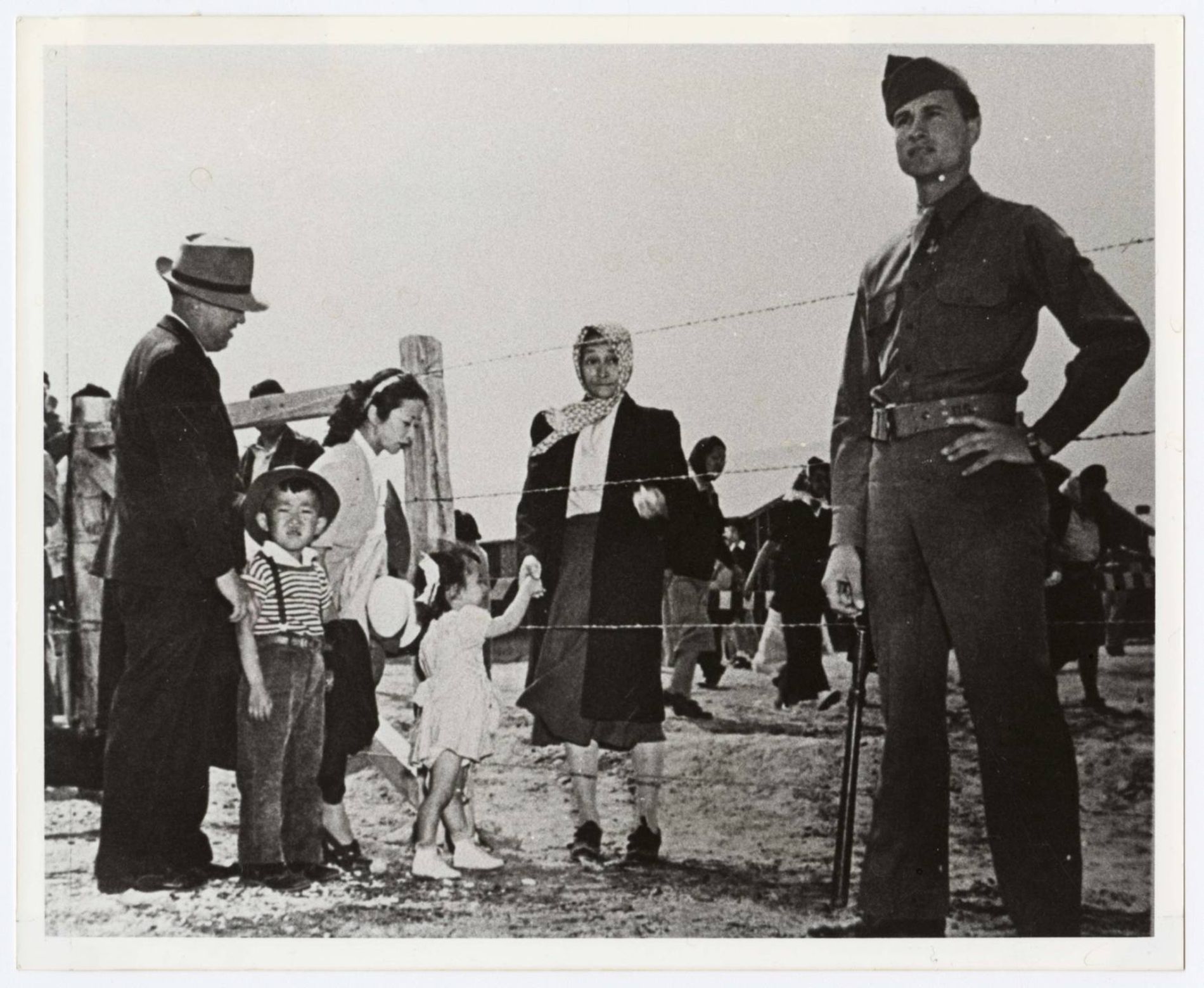 A guard stands next to a family visiting through a barbed wire fence. | Credit: Densho