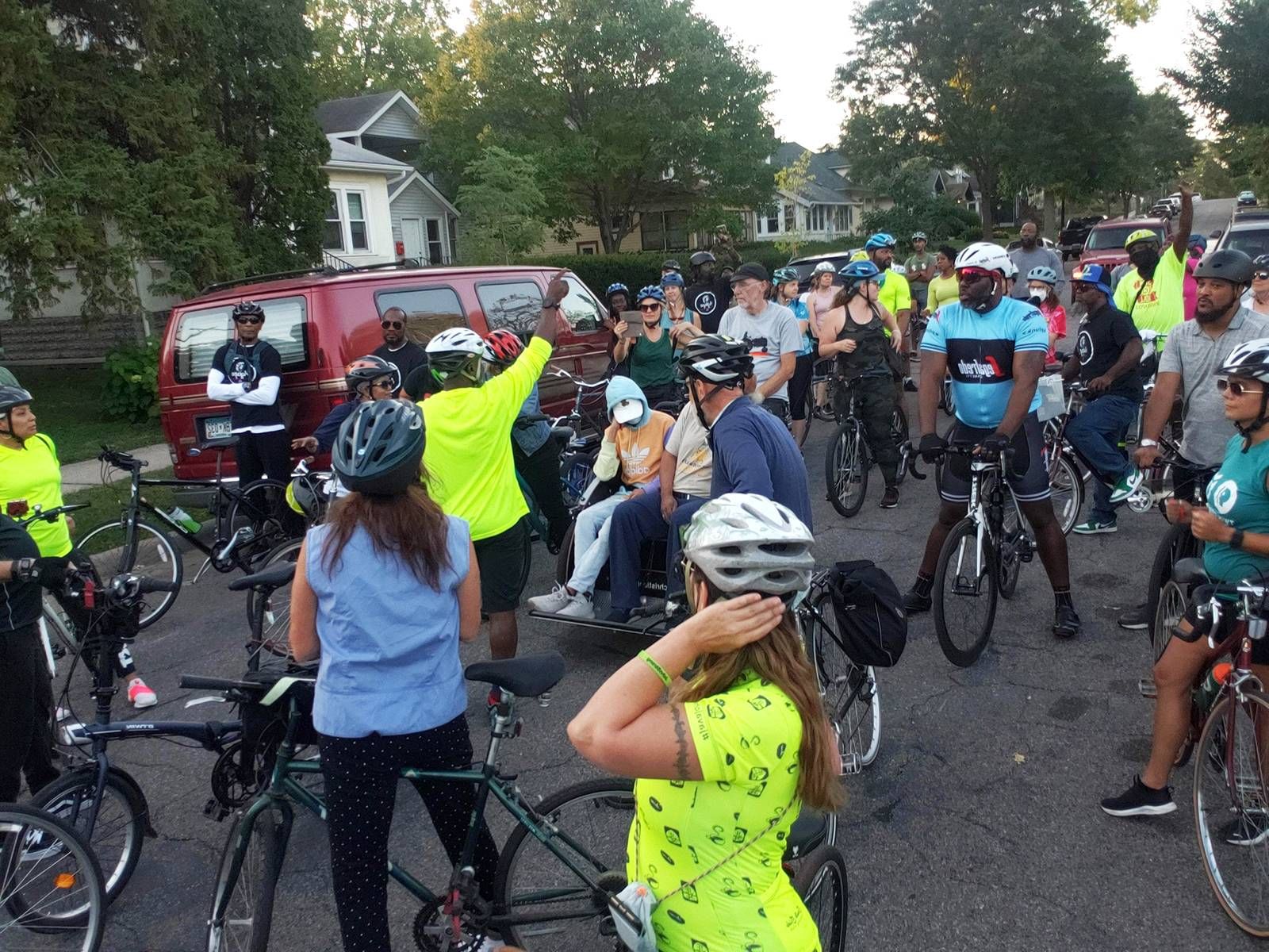 A group of bicyclists stand in a city street, listening to a speaker.