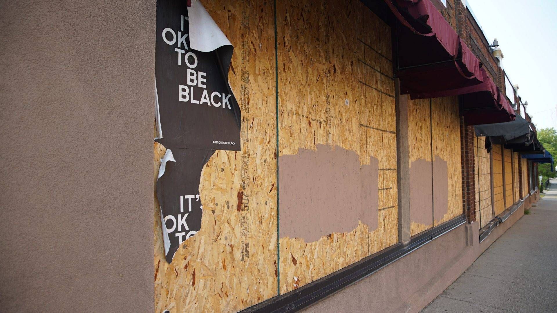 Ripped signs reading "It's OK to be Black" on plywood covering a Minneapolis building.