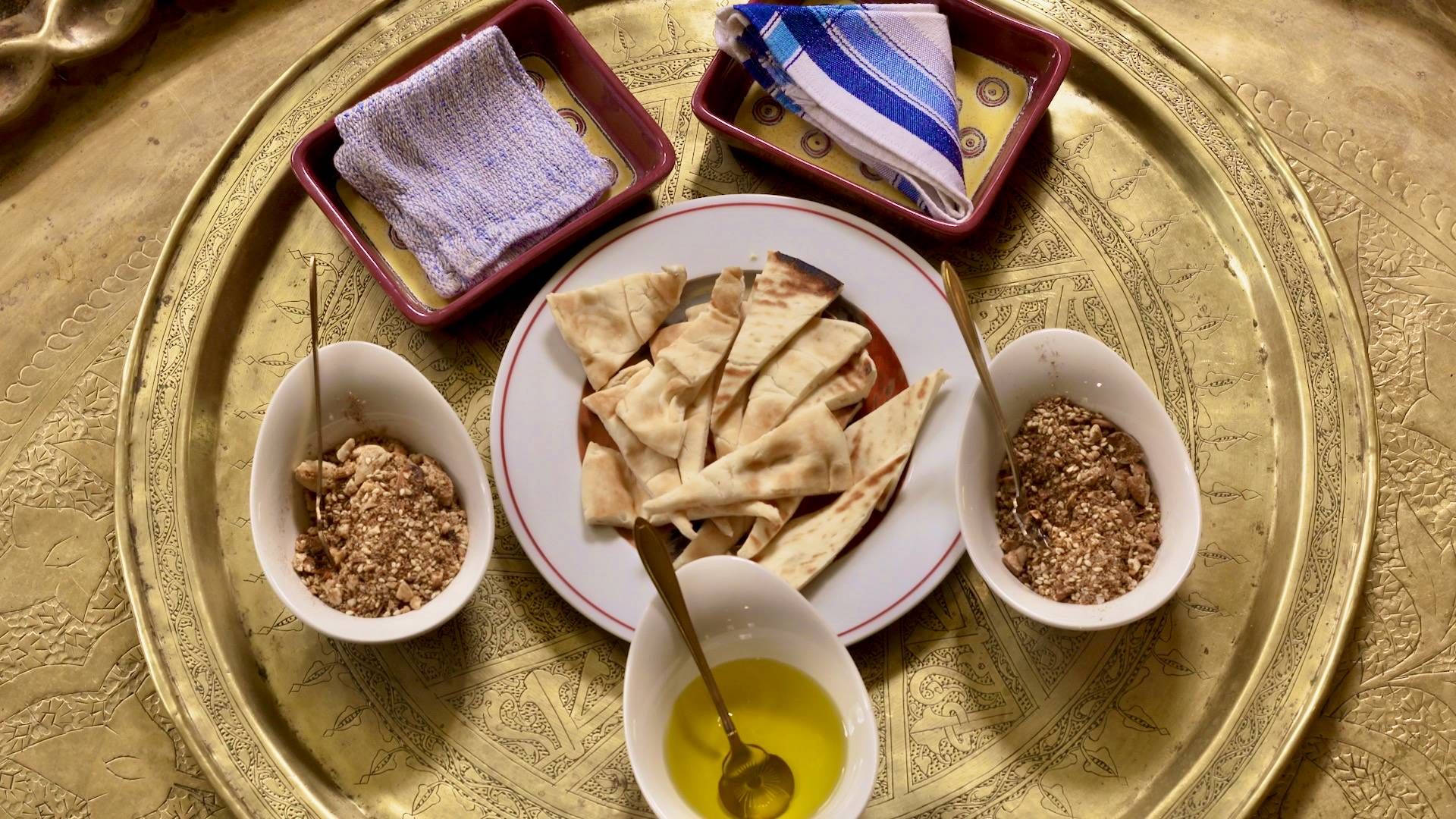 Gold platter with engravings holds two folded napkins, two small bowls of dukkah with serving spoons, a small bowl of olive oil with a serving spoon and a small plate of pita bread.