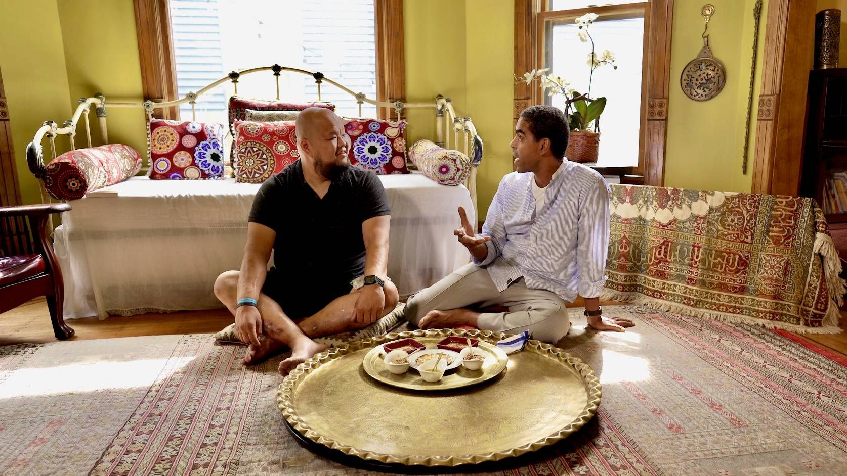 Yia Vang and Mo Kotb sitting on pillows talking around a platter of dukkah, pita and olive oil.
